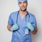 Médecin positif en uniforme bleu et gants de protection debout avec un stéthoscope et regardant la caméra sur fond clair