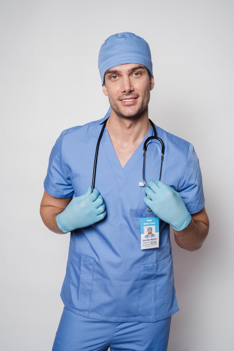 Médecin positif en uniforme bleu et gants de protection debout avec un stéthoscope et regardant la caméra sur fond clair